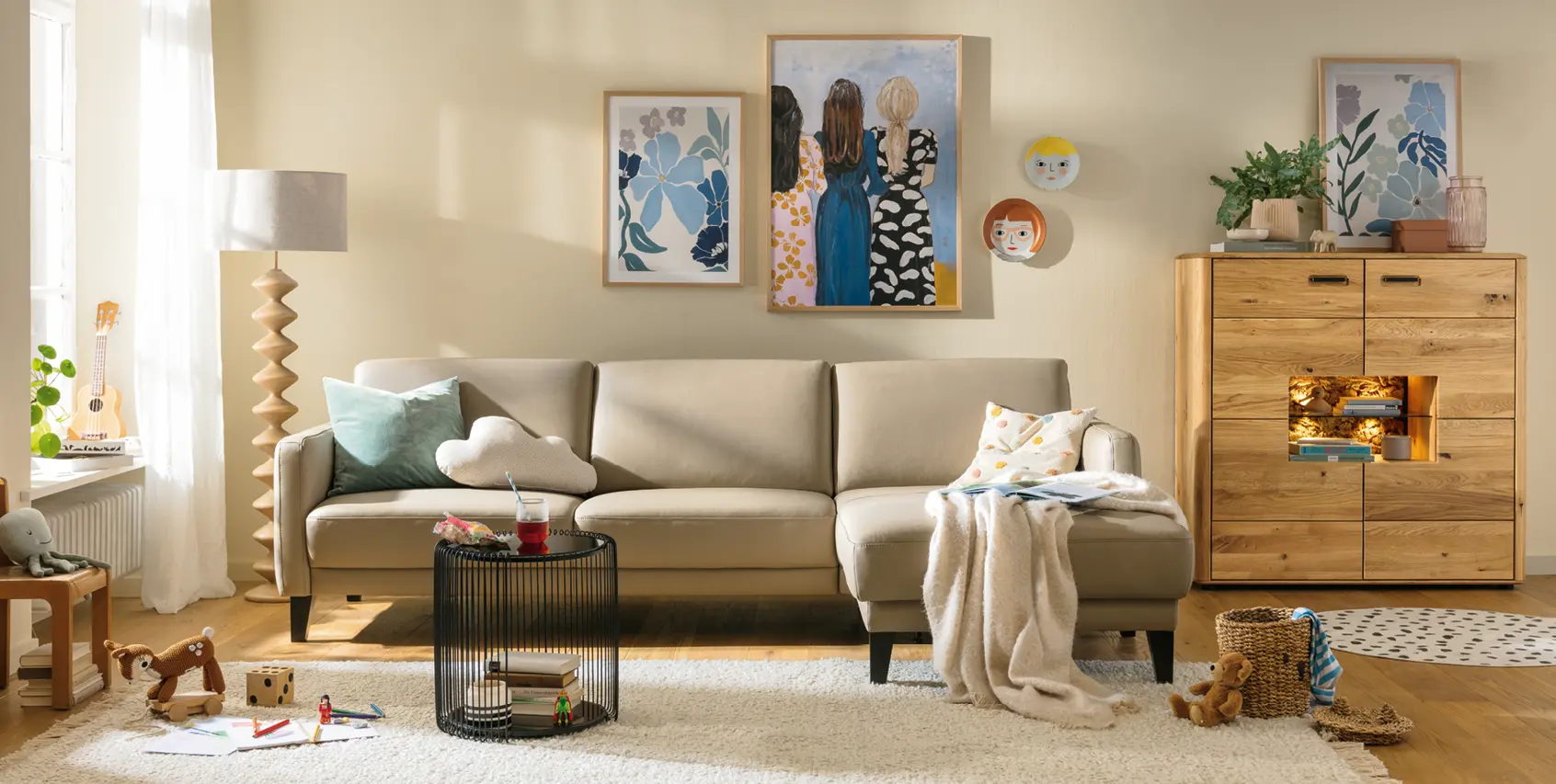 Helles Wohnzimmer mit beigem Ecksofa, Couchtisch und Massivholz-Kommode, dekoriert mit Kissen, Decke und Bildern an der Wand. Gemütliche Wohnidee im modernen Stil mit natürlichen Materialien und warmem Licht.
